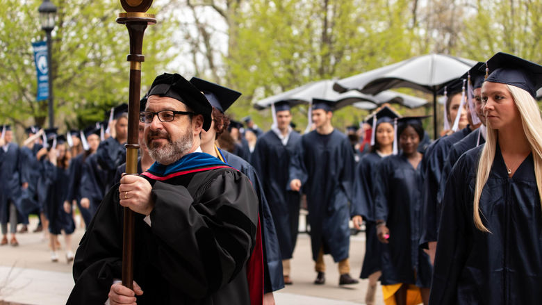 A male professor in a black academic robe carries a mace and leads two rows of graduates in dark blue caps and gowns to the graduation ceremony. 