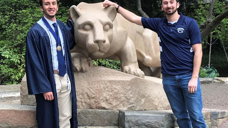Male graduate in cap and gown and another male student standing next to Nittany Lion statue.