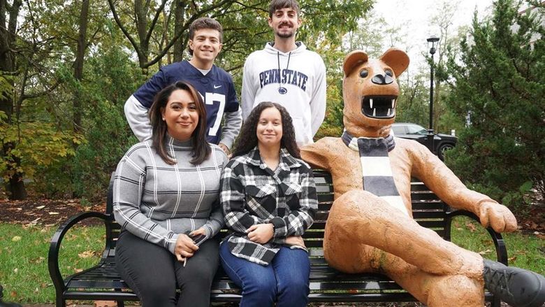 Four students gathered around a bench on a sidewalk seated next to a Nittany Lion statue.