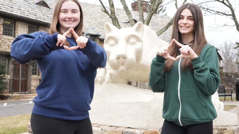 Two female students making diamond shape with their hands in front of a Nittany Lion statue.