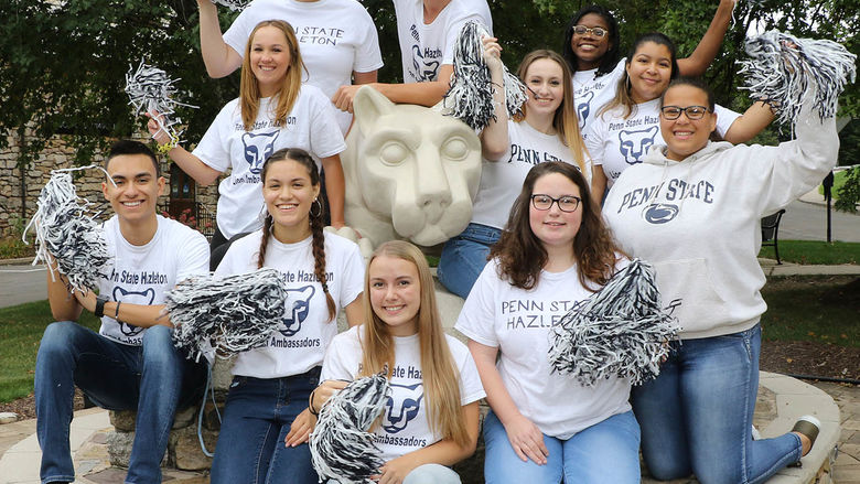 Students in white T-shirts gathered around Nittany Lion statue waving pom-poms.