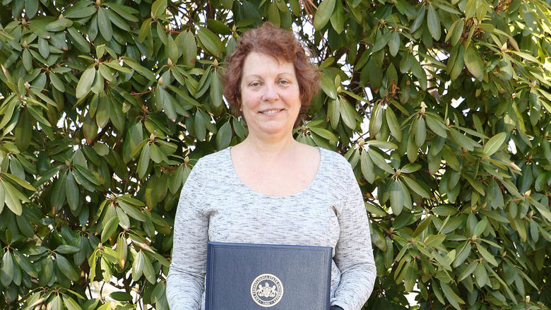 Woman holding plaque in front of bush with green leaves.