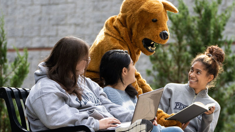 Three female students on a bench outdoors with Nittany Lion mascot leaning over them pointing to a book.