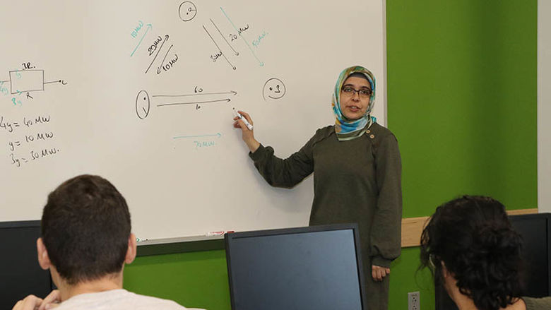 Penn State Hazleton Assistant Teaching Professor Mesude Bayrakci-Boz standing in front of a white board teaching with students in desks looking on. 