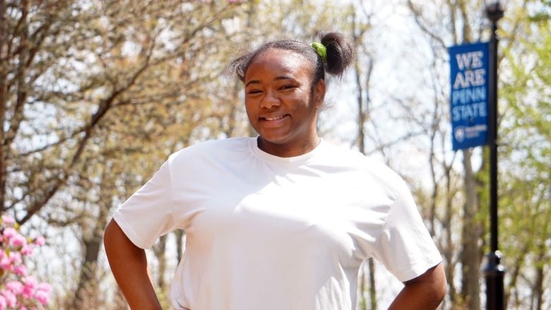 Female student in white T-shirt standing on sidewalk surrounded by trees.