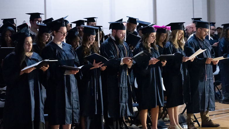 Line of graduates reading from a booklet while standing.