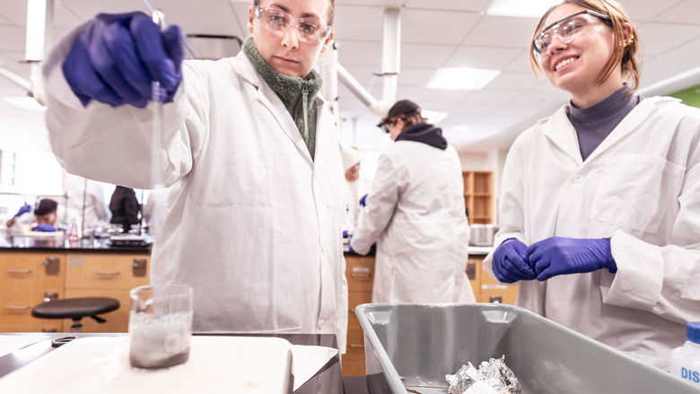 Two students in white lab coats in a chemistry laboratory mixing chemicals in a glass beaker.