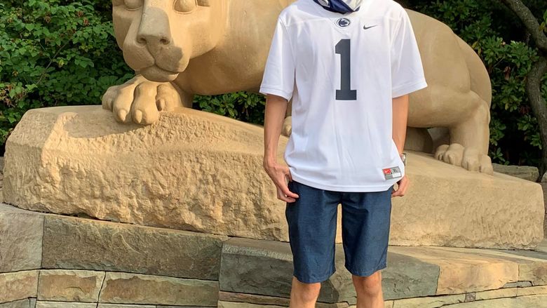 Student in Penn State football jersey standing in front of Nittany Lion statue