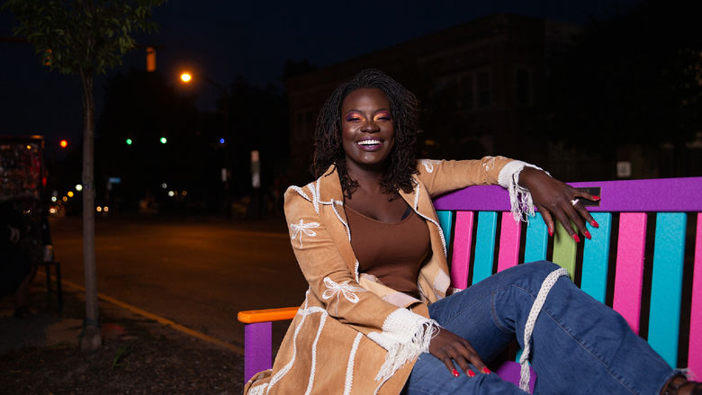Woman in tan jacket sitting on multicolored park bench at night.