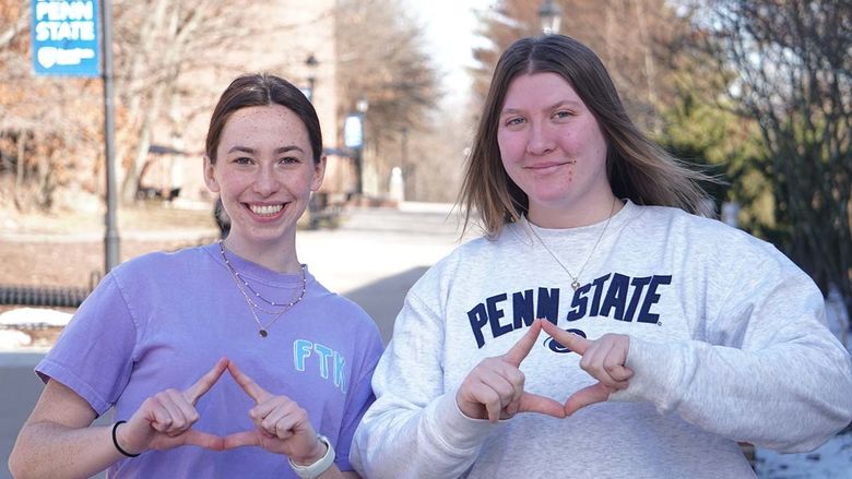 Two female students on campus mall making diamond symbol with their hands.