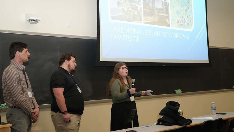 Three students standing beside projector screen in front of classroom.
