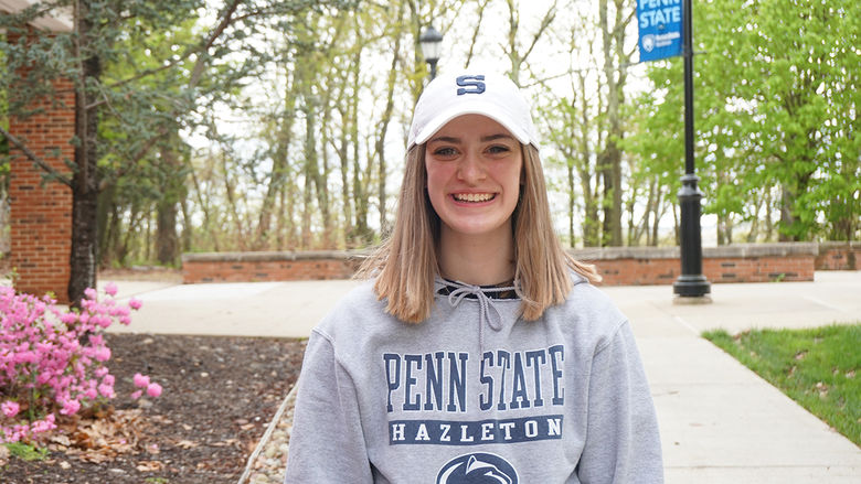 Student in white Penn State hat and gray Penn State Hazleton hoodie standing outside on campus mall.