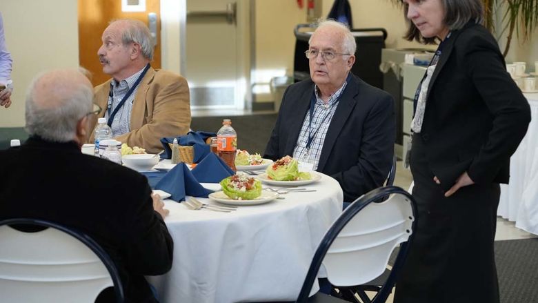 Group of men sitting at a table with woman standing next to them speaking.