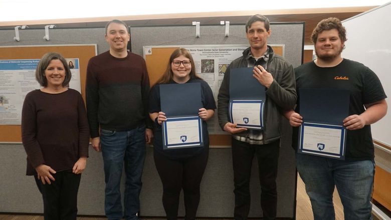 Four students holding certificates in front of poster board.