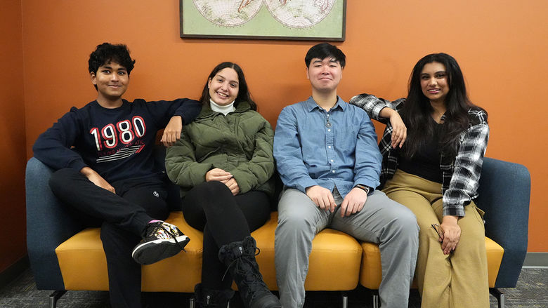 Four students seated next to one another on a couch up against an orange wall.