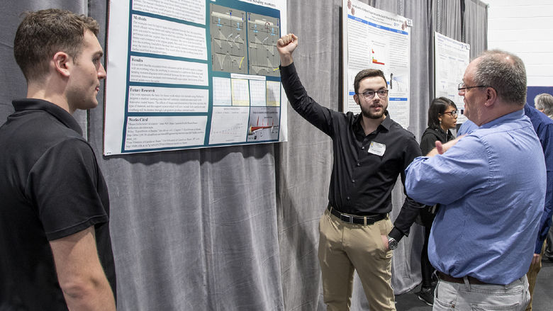 Student wearing black shirt points to research poster as he speaks with a symposium judge.