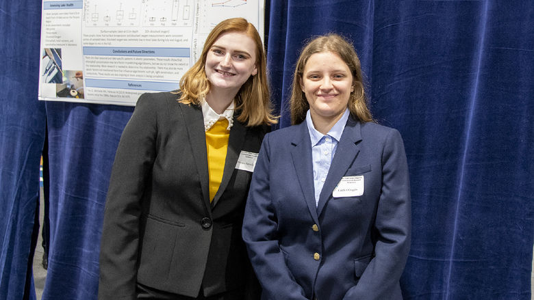 Two students wearing suits pose for a photo in front of a research poster.
