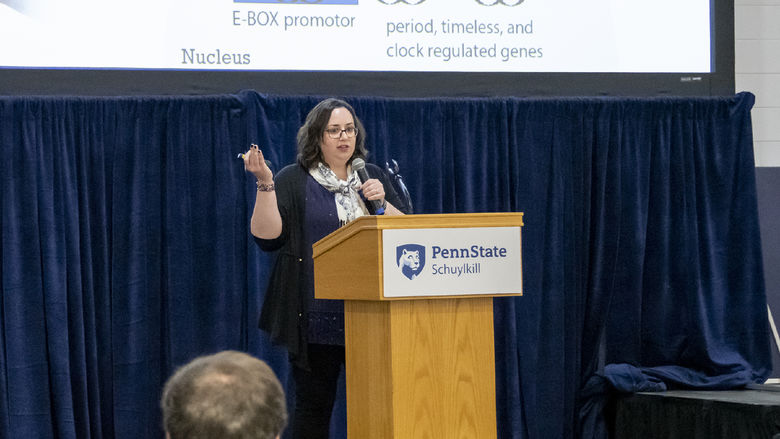 Woman stands in front of a projector screen and behind a podium, holding a microphone, as she gestures to her PowerPoint slide