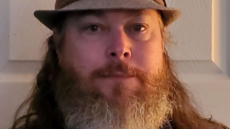Man with a grey beard wearing tan fedora-style hat.
