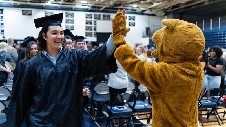 A graduate in a blue cap and gown high fives a Nittany Lion mascot.