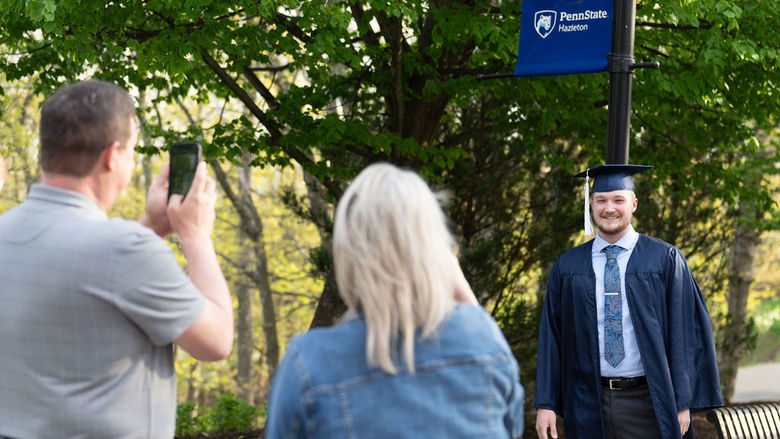 Student in cap and gown having photo taken by man and woman in front of a tree and light post.