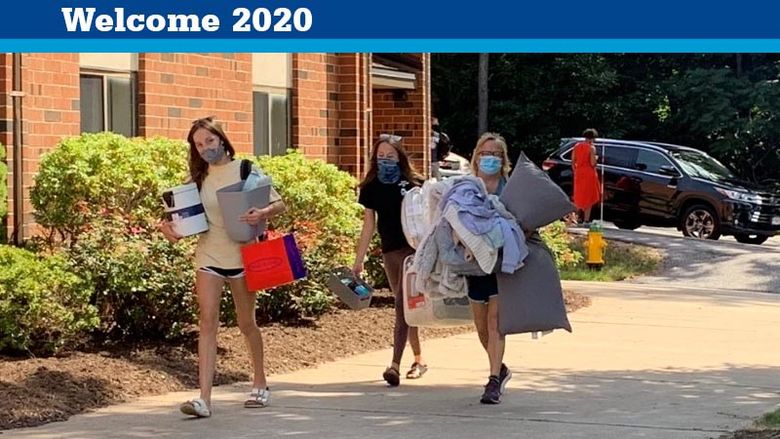 Three females wearing masks and carrying items into a residence hall. 