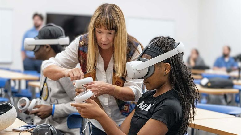Female student wearing white virtual reality headset with female professor standing next to her offering guidance,