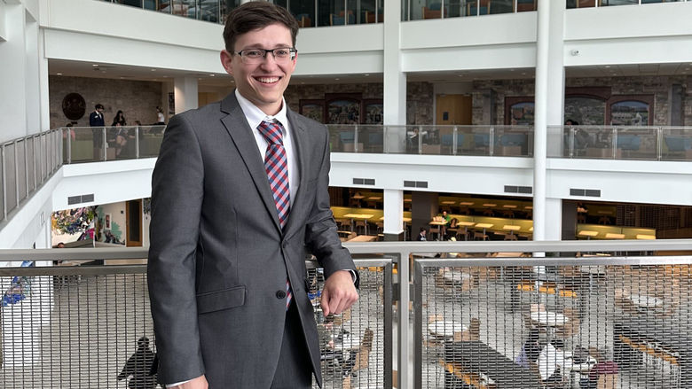 Male student in gray suit standing against a fenced balcony overlooking a caferteria