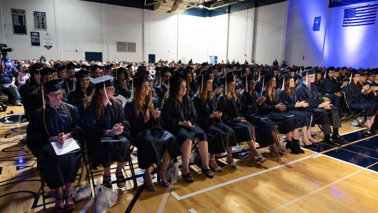 Students in caps and gowns seated on a hardwood gym floor.