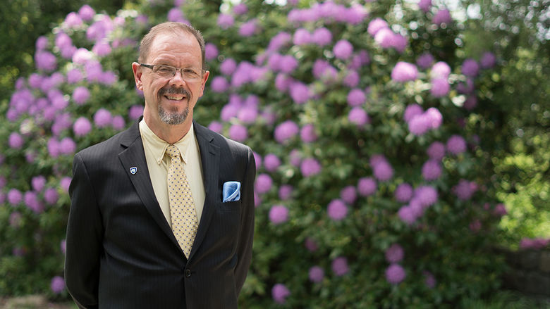 Man in a suit standing in front of a bush with purple flowers.
