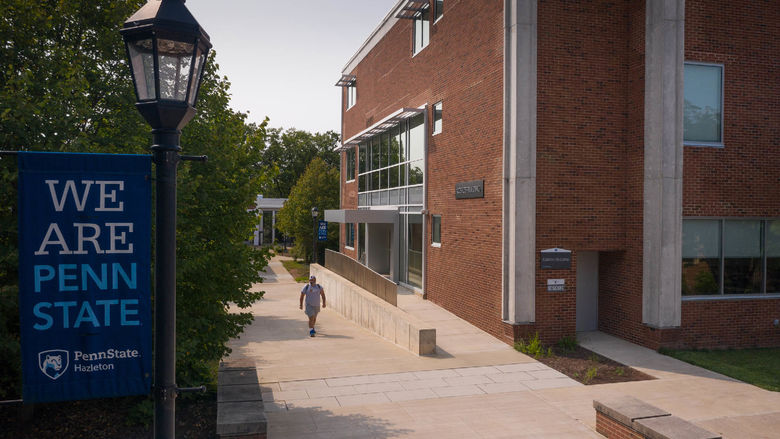 Student walking along sidewalk in between trees and a large brick building.
