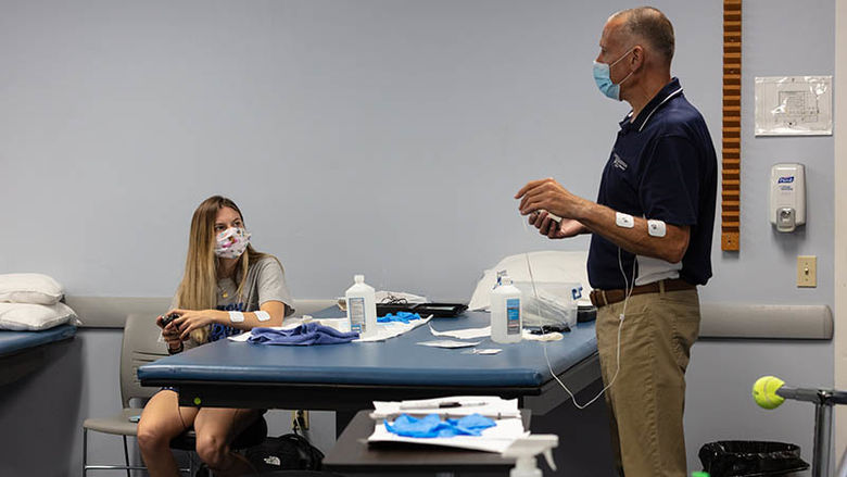 A male instructor demonstrates a TENS unit on his arm as a female student watches. 