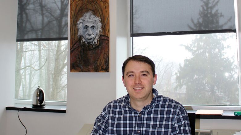 Joe Ranalli in collared shirt sitting at office desk with Albert Einstein poster in background