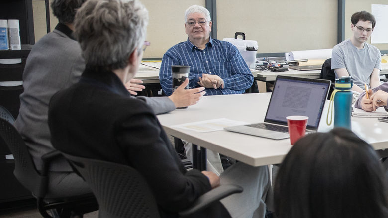 A man sitting at a table listening to someone on his right speak as others at the table look on.