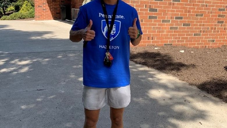 Student wearing mask and blue Penn State Hazleton shirt giving two thumbs up outside North Hall dorm.