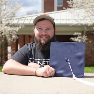 Man leaning over wall holding graduation cap in left hand and smiling.