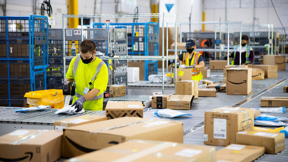Amazon employees wearing masks sort numerous packages at a fulfillment center.