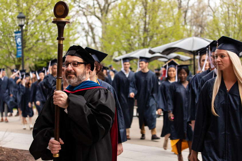 A male professor in a black academic robe carries a mace and leads two rows of graduates in dark blue caps and gowns to the graduation ceremony. 