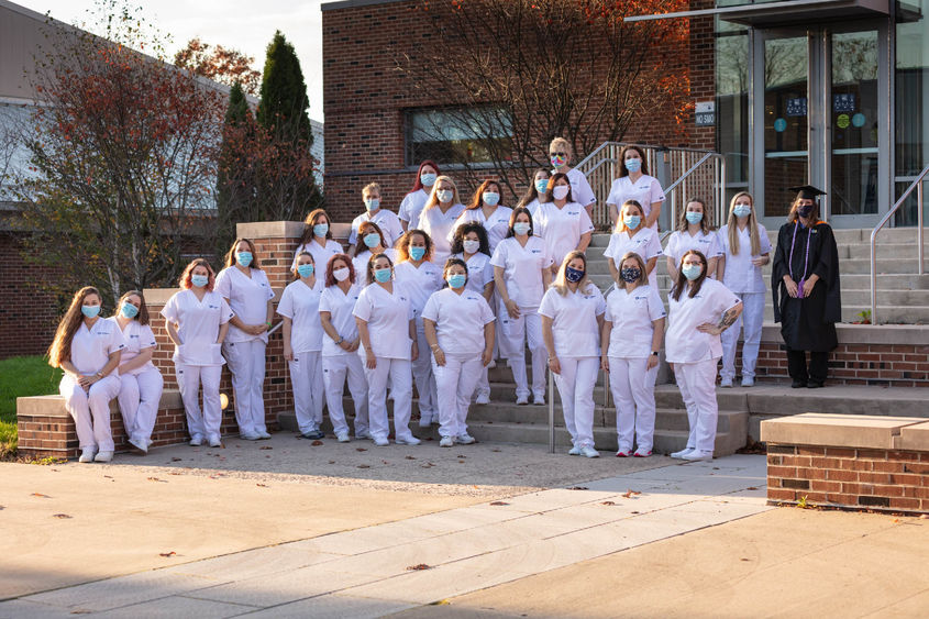 Rows of practical nursing students in white scrubs lined up on steps in front of campus building.