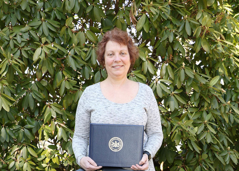 Woman holding plaque in front of bush with green leaves.