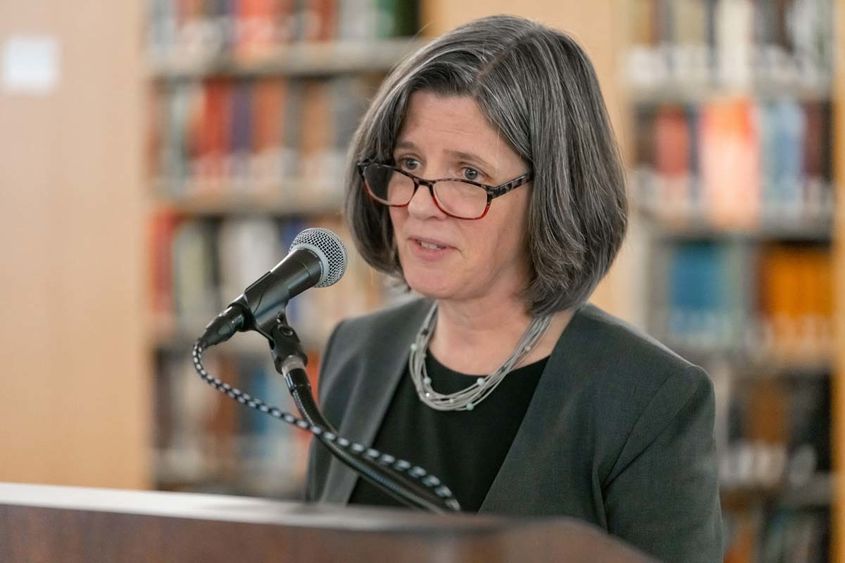 A woman in glasses smiles from behind a microphone at a podium,