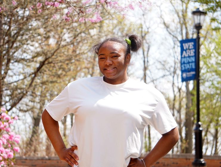 Female student in white T-shirt standing on sidewalk surrounded by trees.