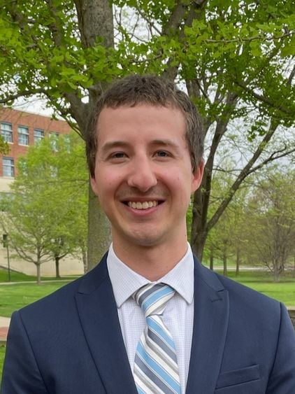 Man in suit and tie standing outdoors in front of a tree.