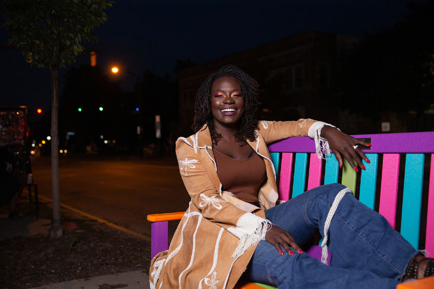 Woman in tan jacket sitting on multicolored park bench at night.