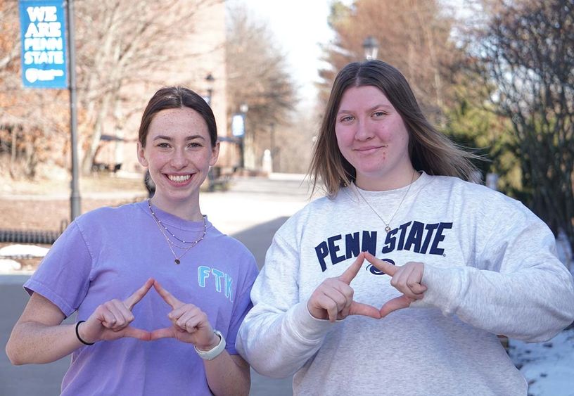 Two female students on campus mall making diamond symbol with their hands.