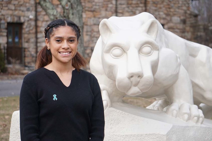 Student with dark hair in a black blouse standing in front of white Nittany Lion statue.