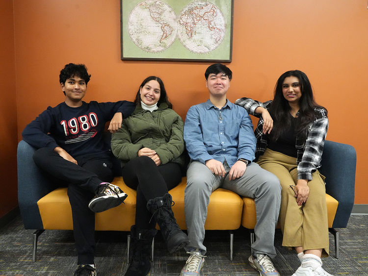 Four students seated next to one another on a couch up against an orange wall.