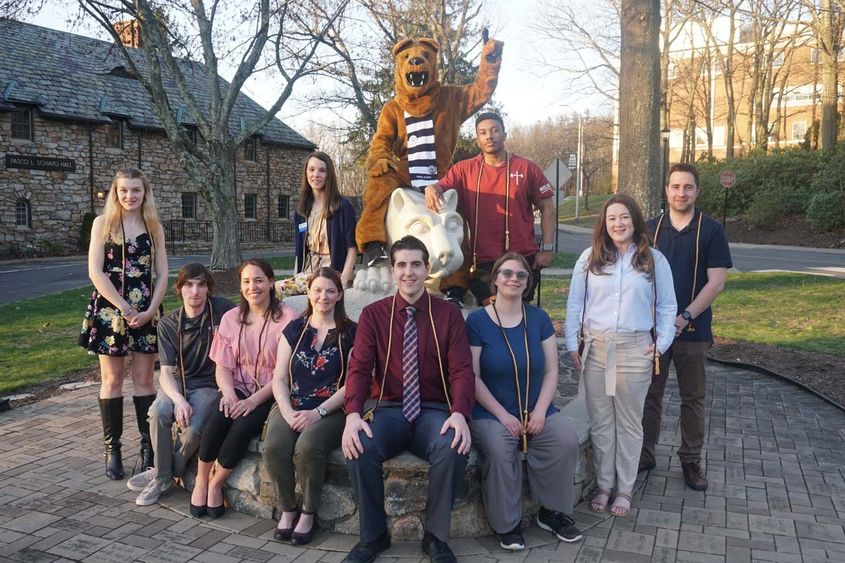 Group of students seated surrounding statue of Nittany Lion in grass field.