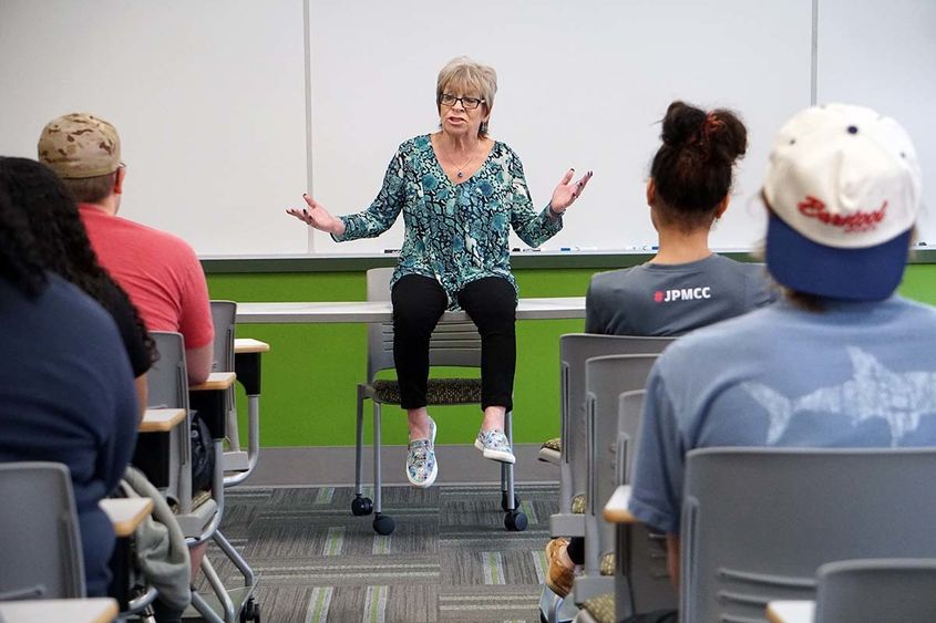 Woman seated on classroom table with rows of students on either side facing her.