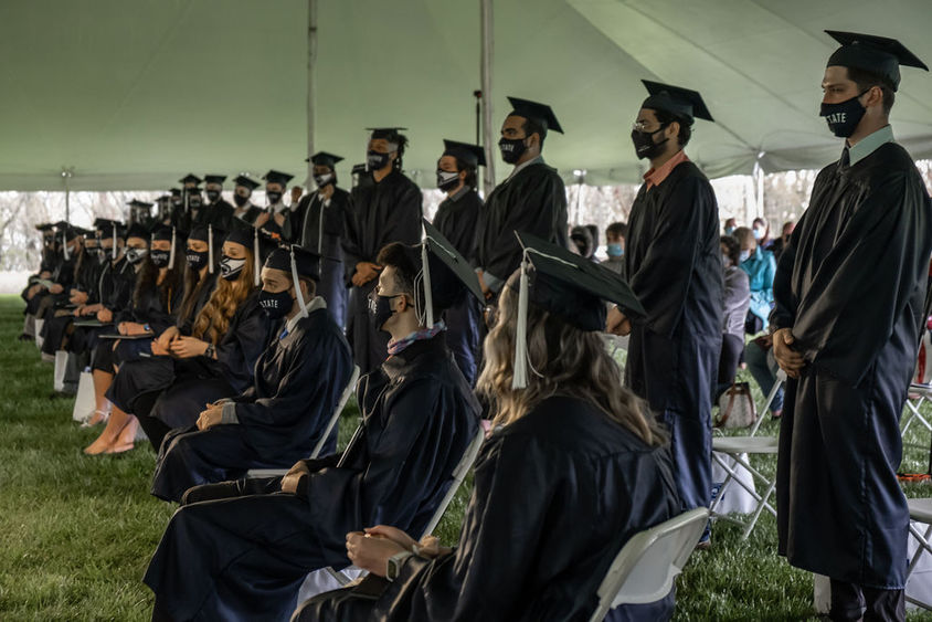 Row of graduates in caps and gowns seated in front of another row of graduates standing.